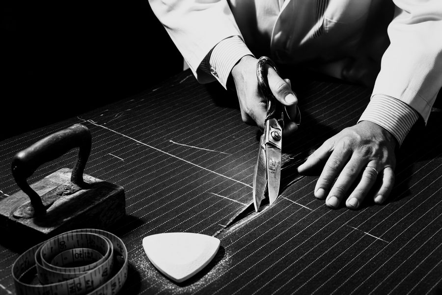 A cutter cuts pinstripe cloth with heavy shears along a chalk line, a tailor's chalk and tape measure beside him.